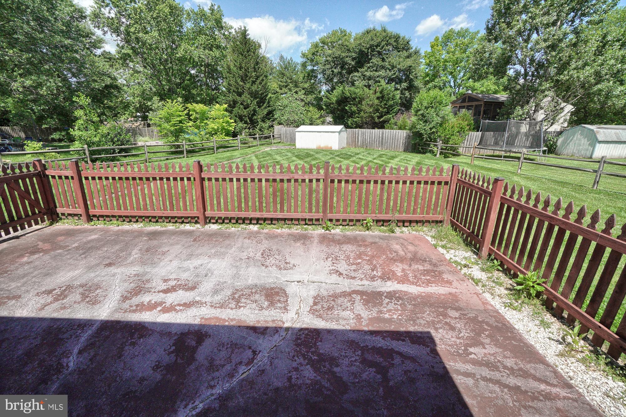 Concrete patio overlooking a fenced, tree-lined backyard with lawn and storage sheds.