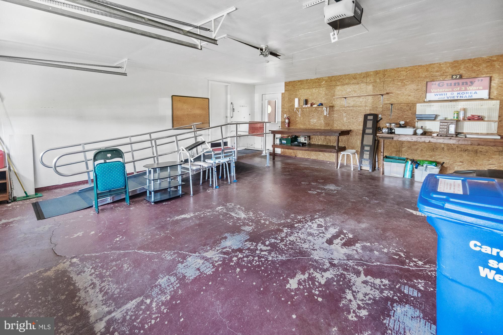 Garage with a metal accessibility ramp and handrails, workbench, plywood wall, and blue recycling bin.