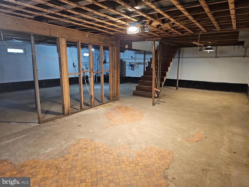 Unfinished basement with exposed ceiling framing, patched concrete floor, and stairs.