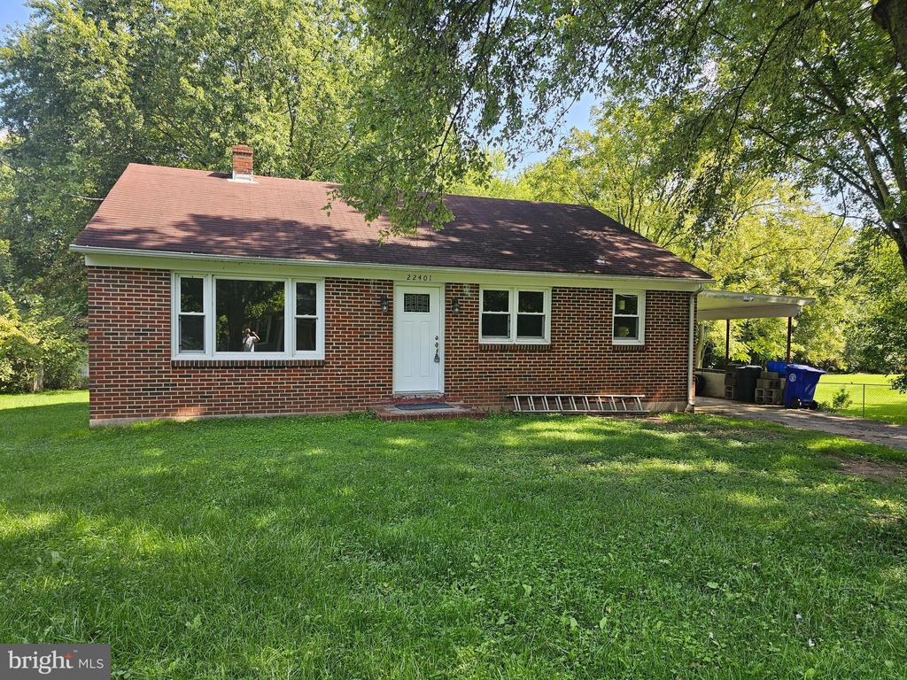 Single-story brick house with a white front door and carport beneath mature trees.