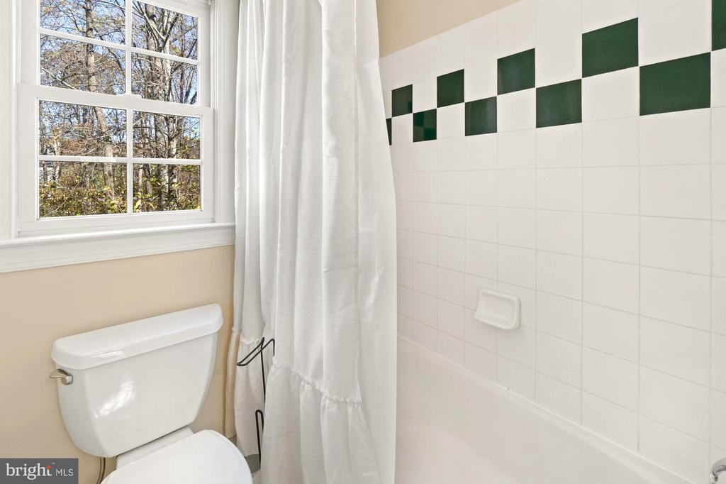 Bathroom with a white-tiled tub and shower, green accent tiles, and a window overlooking trees.