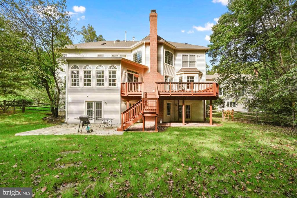 Rear of a two-story house with an elevated deck, brick chimney, and tree-lined lawn.