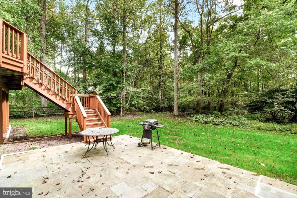 Stone patio with a raised wooden deck, outdoor table, and grill beside a wooded yard.