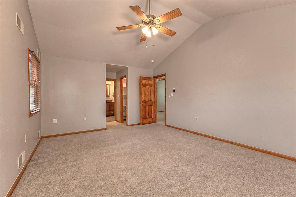 Empty carpeted bedroom with a ceiling fan, wooden window blinds, and open doorways.