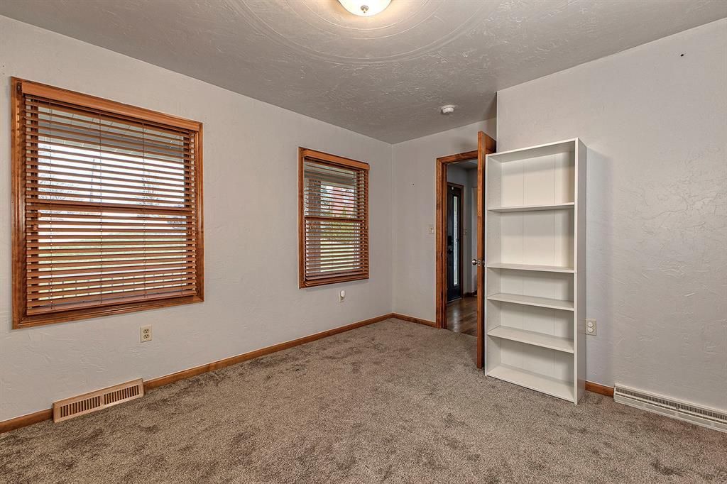 Empty carpeted room with two wood-blind windows and a white shelving unit.
