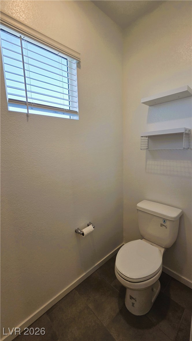 Bathroom with a white toilet, dark tile floor, high window with blinds, and wall-mounted shelves.