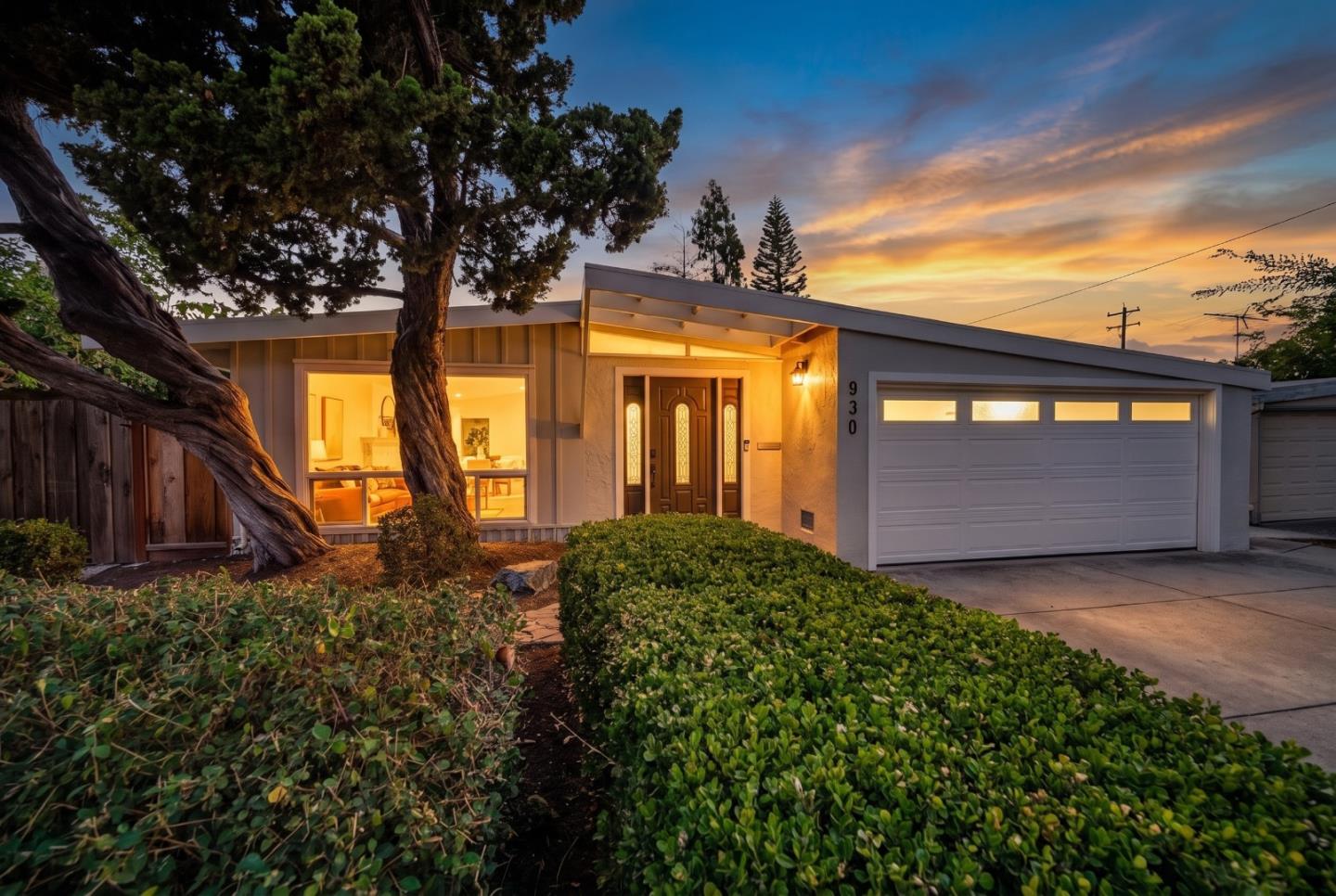Modern single-story home at dusk with a warmly lit entry, broad garage, and mature trees.