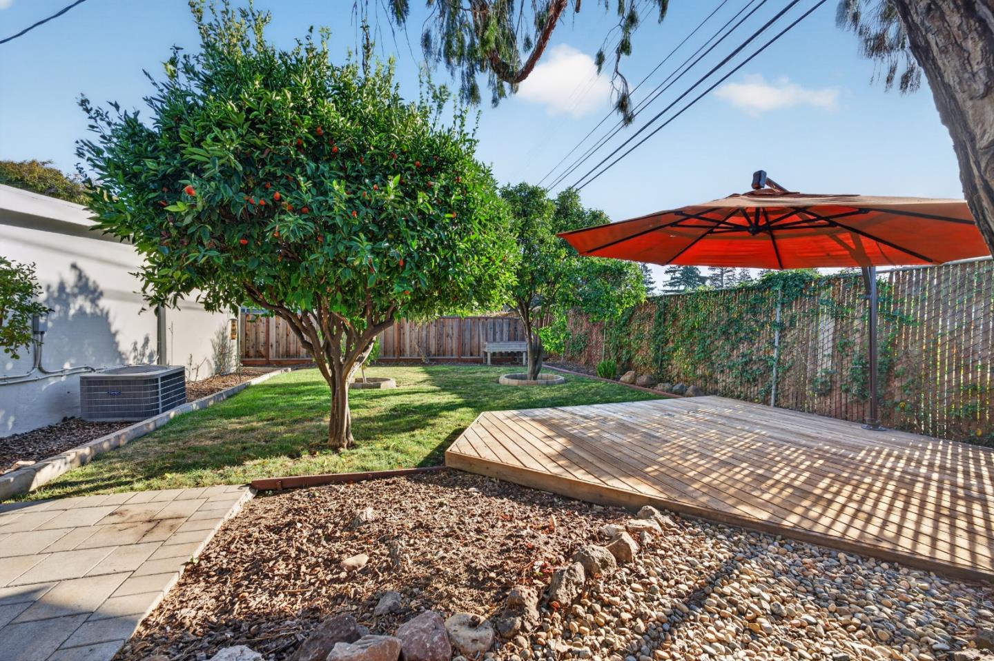 Fenced backyard with a raised wooden deck, red umbrella, mature tree, and stone landscaping.