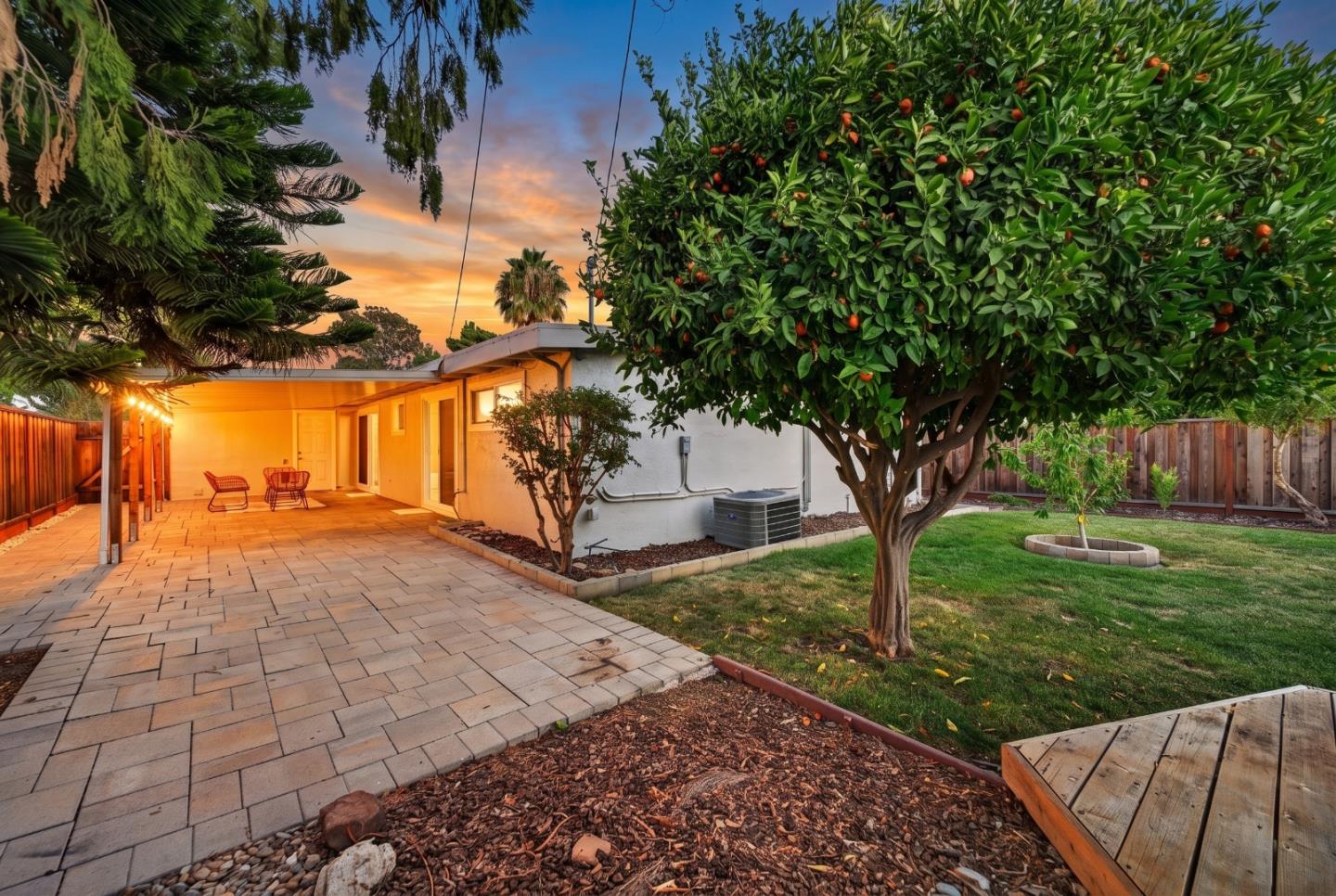 Backyard patio with a covered seating area, paver walkway, lawn, and fruiting citrus tree at dusk.