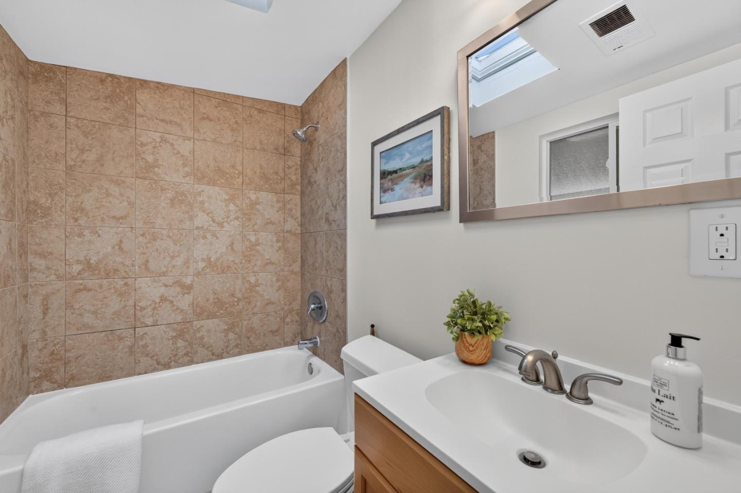 Bathroom with tan tiled tub surround, wood vanity, and skylight reflected in the mirror.