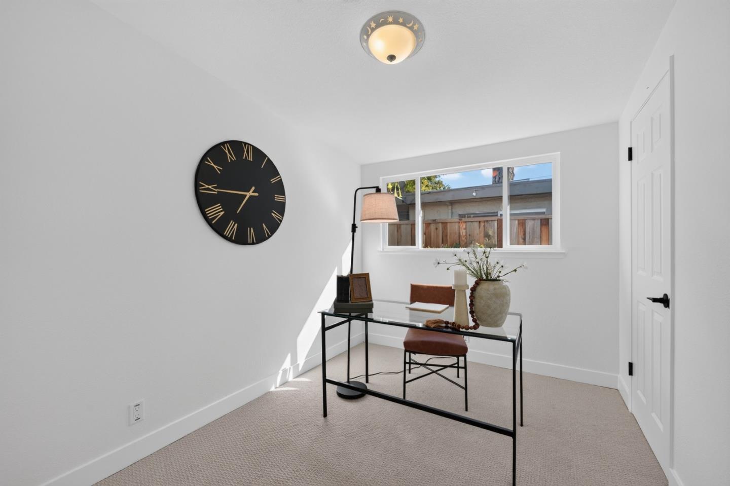 Staged home office with a glass desk, brown chair, large wall clock, and window overlooking a wooden fence.