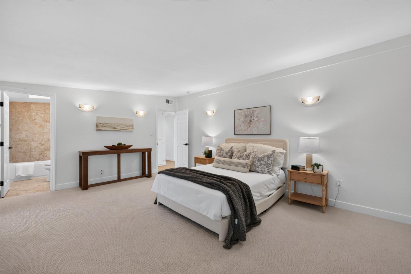 Staged bedroom with an upholstered bed, dark knitted throw, beige carpet, and adjoining tiled bathroom.