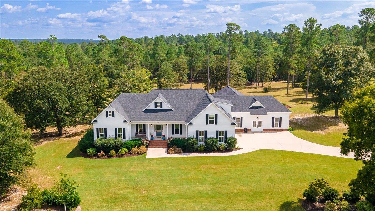 Aerial view of a white house with a broad lawn, curved driveway, and wooded surroundings.
