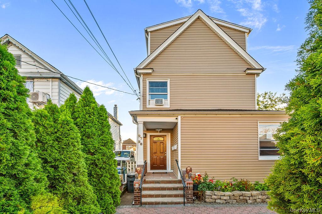 Beige two-story house with a steep gabled roof, brick entry steps, and tall evergreen hedges.