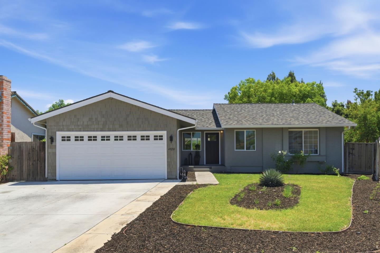 Gray single-story house with a white double garage door, concrete driveway, and landscaped front lawn.