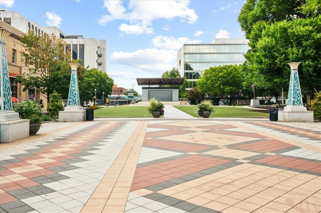 Public plaza with patterned paving, a lawn, mature trees, and modern buildings.