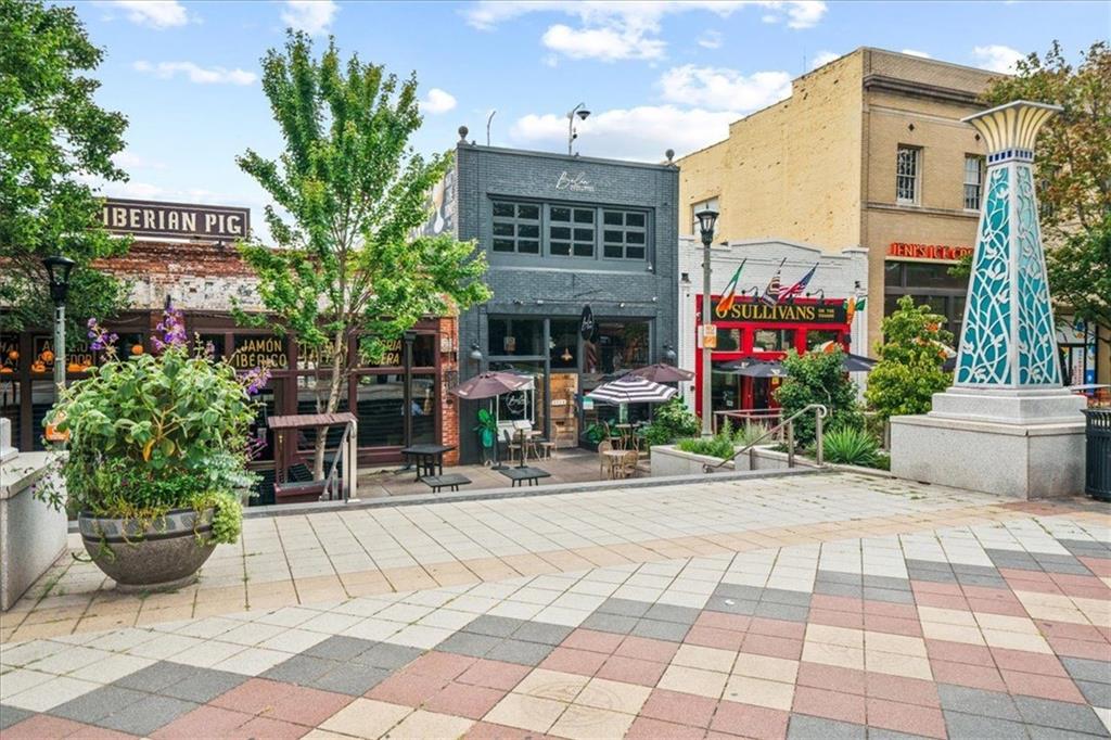 Pedestrian plaza with restaurant storefronts, outdoor tables, colorful paving, and a tiled monument.