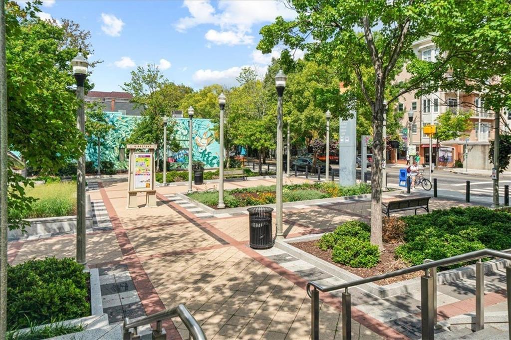 Tree-shaded public plaza with patterned paths, benches, flower beds, and a colorful mural.
