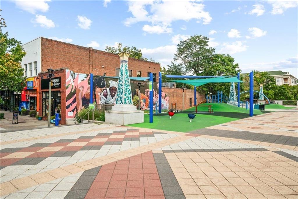 Neighborhood plaza with a geometric tiled walkway, mural-covered storefront, and shaded green play area.