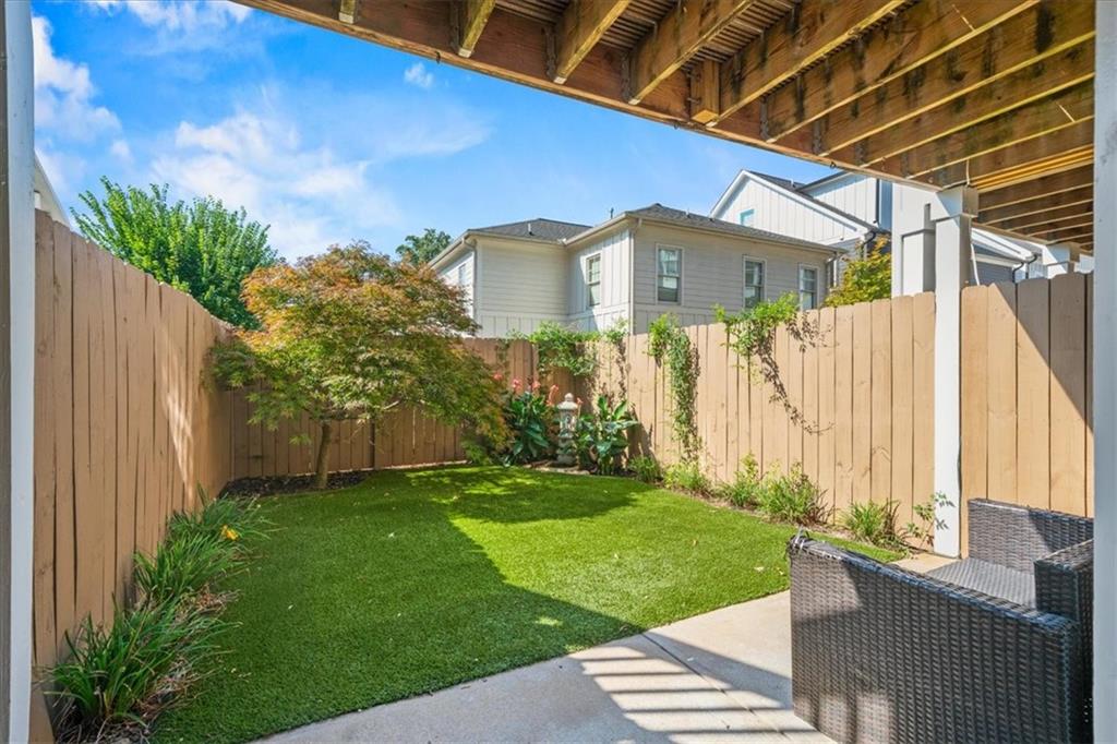 Fenced backyard patio with a green lawn, leafy landscaping, and wicker seating beneath exposed wood beams.