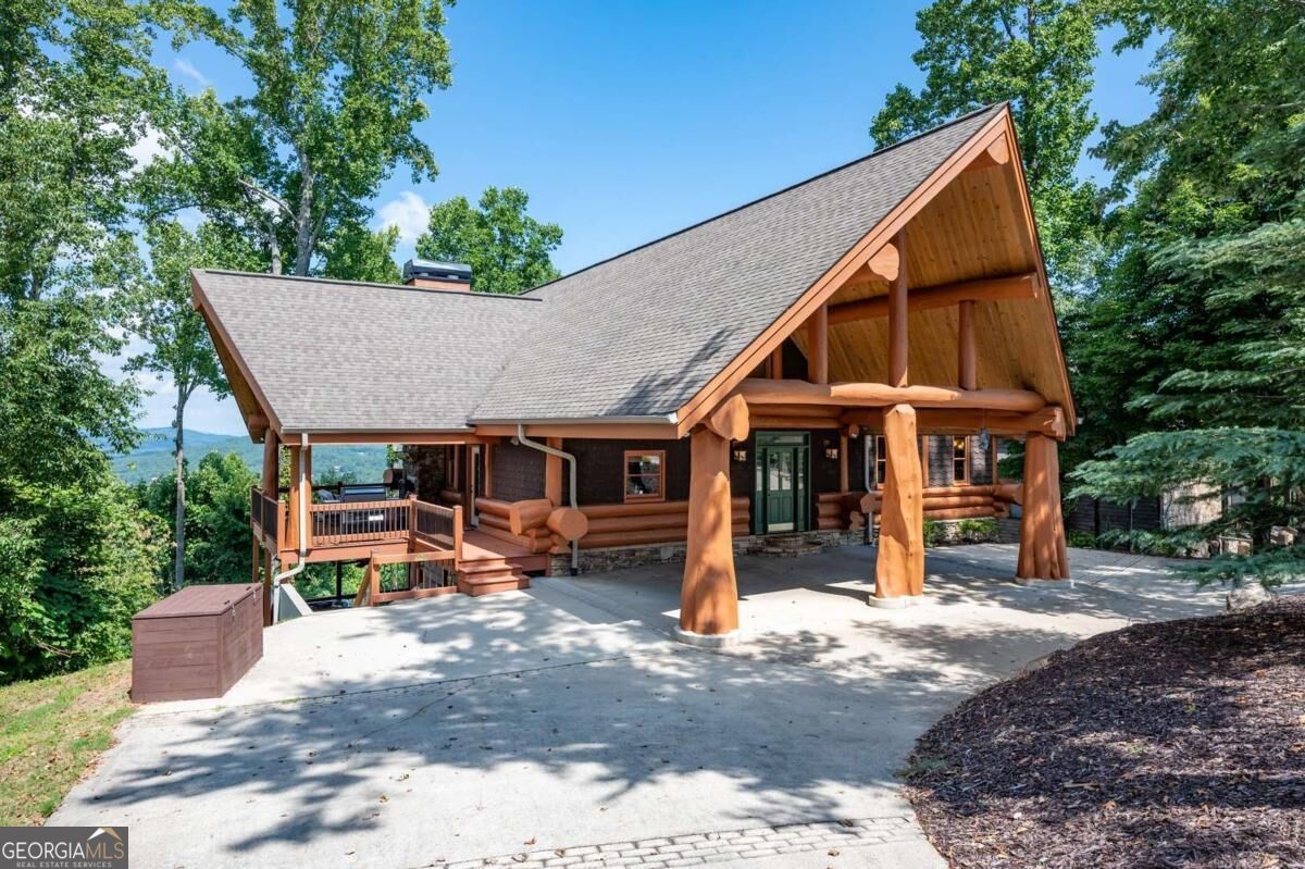 Log cabin with a covered porch and broad concrete driveway, surrounded by trees with distant hills.
