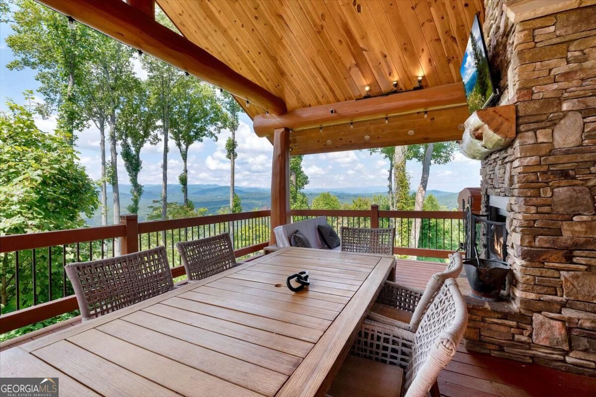 Covered wooden deck with a dining table and stone fireplace overlooking wooded mountains.