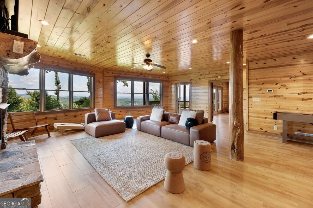 Wood-paneled living room with a stone fireplace, sectional sofa, and windows overlooking wooded hills.