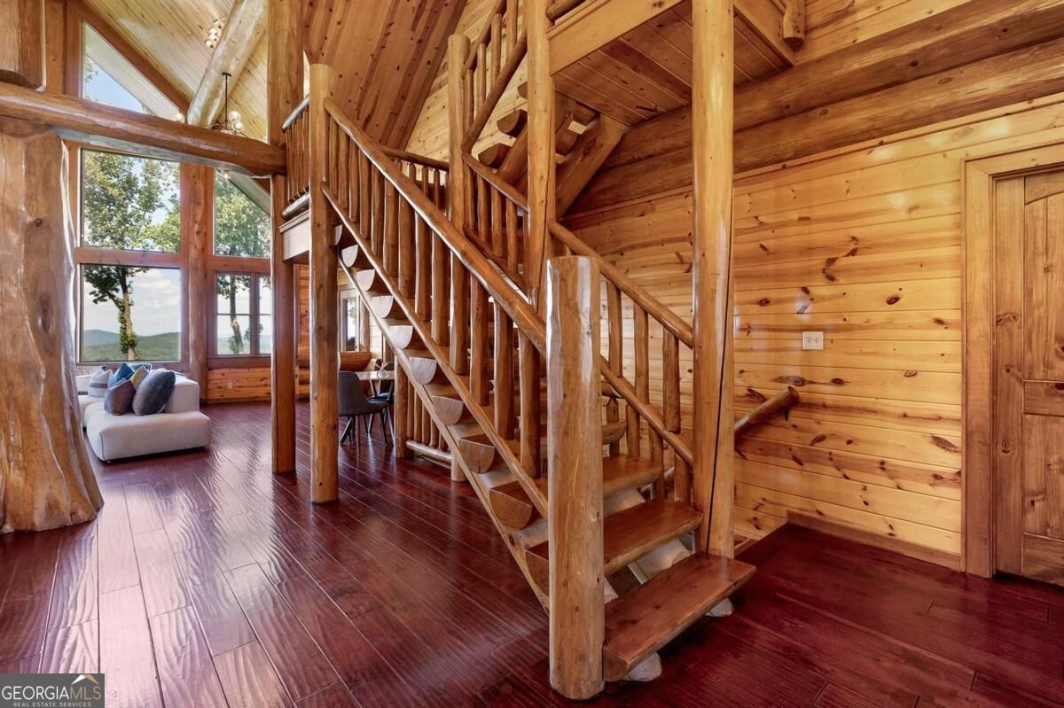 Cabin interior with an open timber staircase, vaulted wood ceiling, and tall windows beside a sofa.
