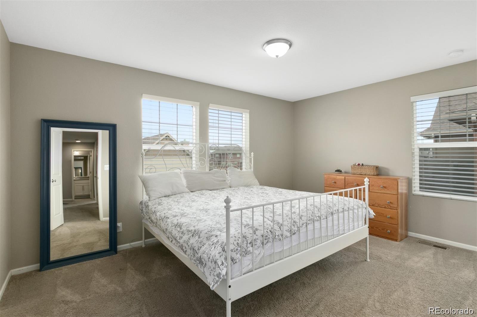 Bedroom with a white metal bed, floral bedding, beige carpet, and windows overlooking neighboring homes.