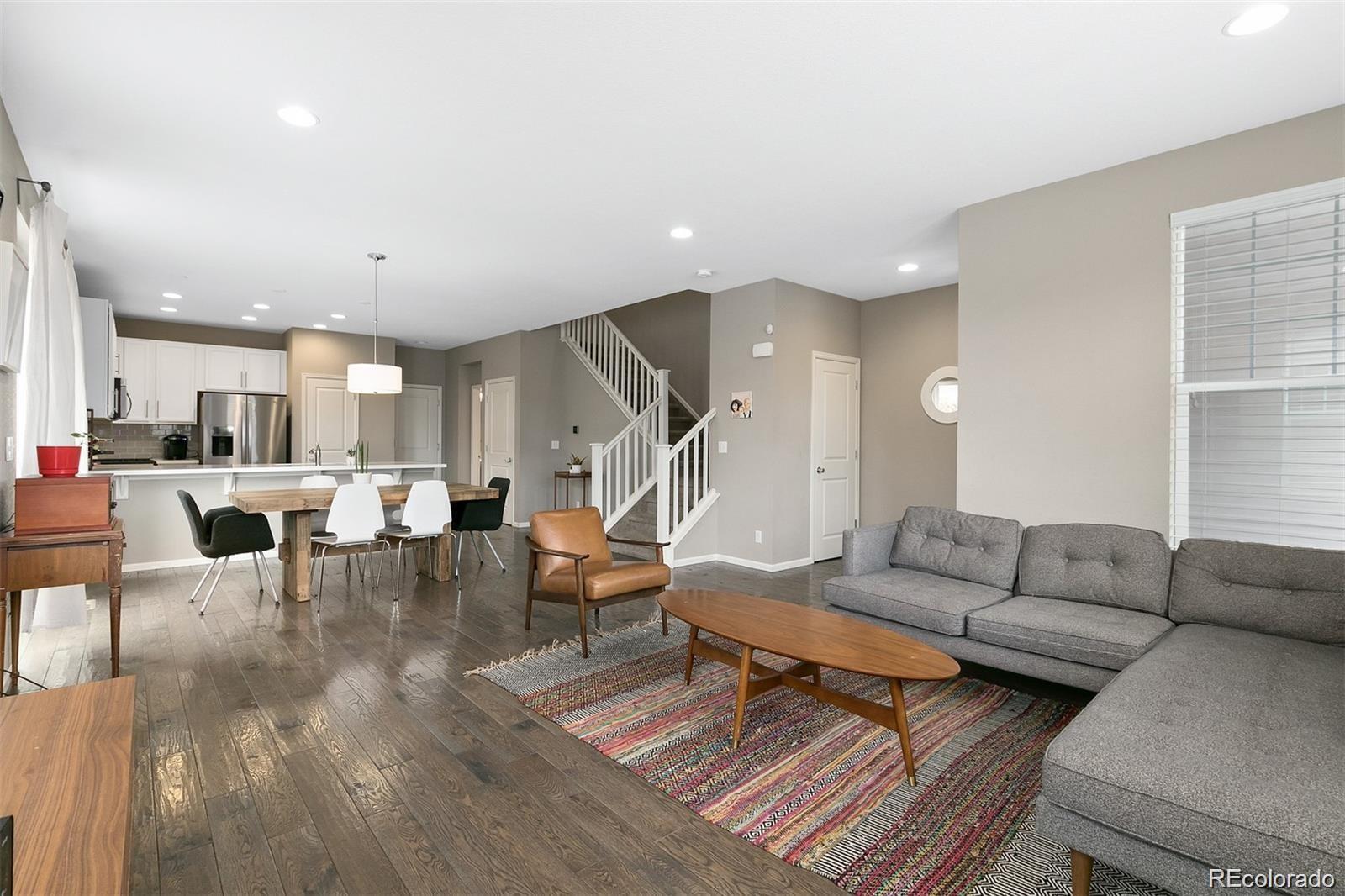 Open-plan living room with gray sectional, colorful rug, dining area, kitchen, and staircase.