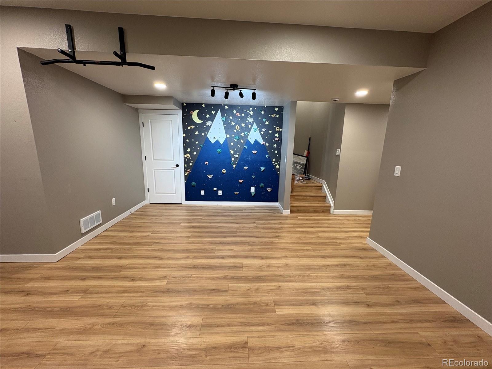 Basement recreation room with a blue climbing wall, wood-look flooring, and stairs.