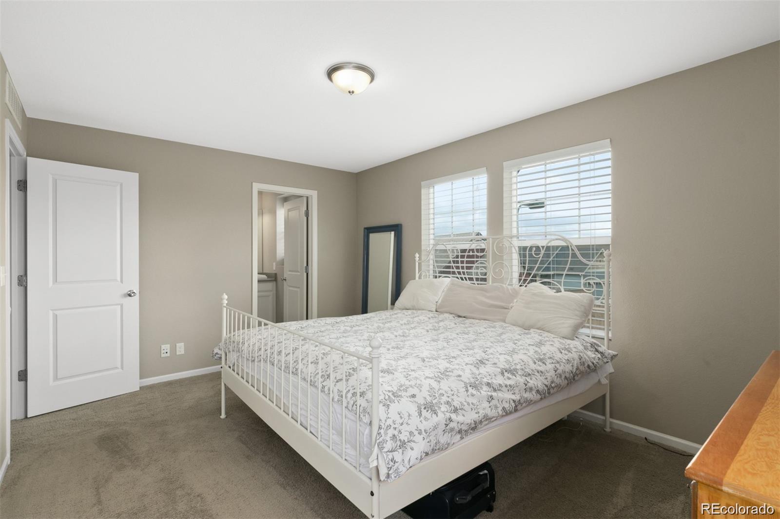 Bedroom with a white metal bed and floral bedding beneath two windows with blinds.