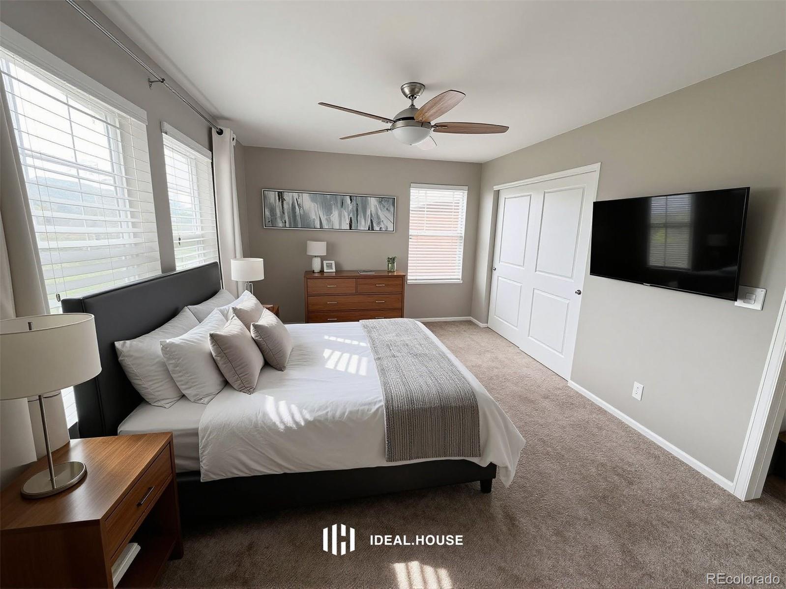 Staged bedroom with a large bed, gray headboard, wood furniture, ceiling fan, and wall-mounted television.
