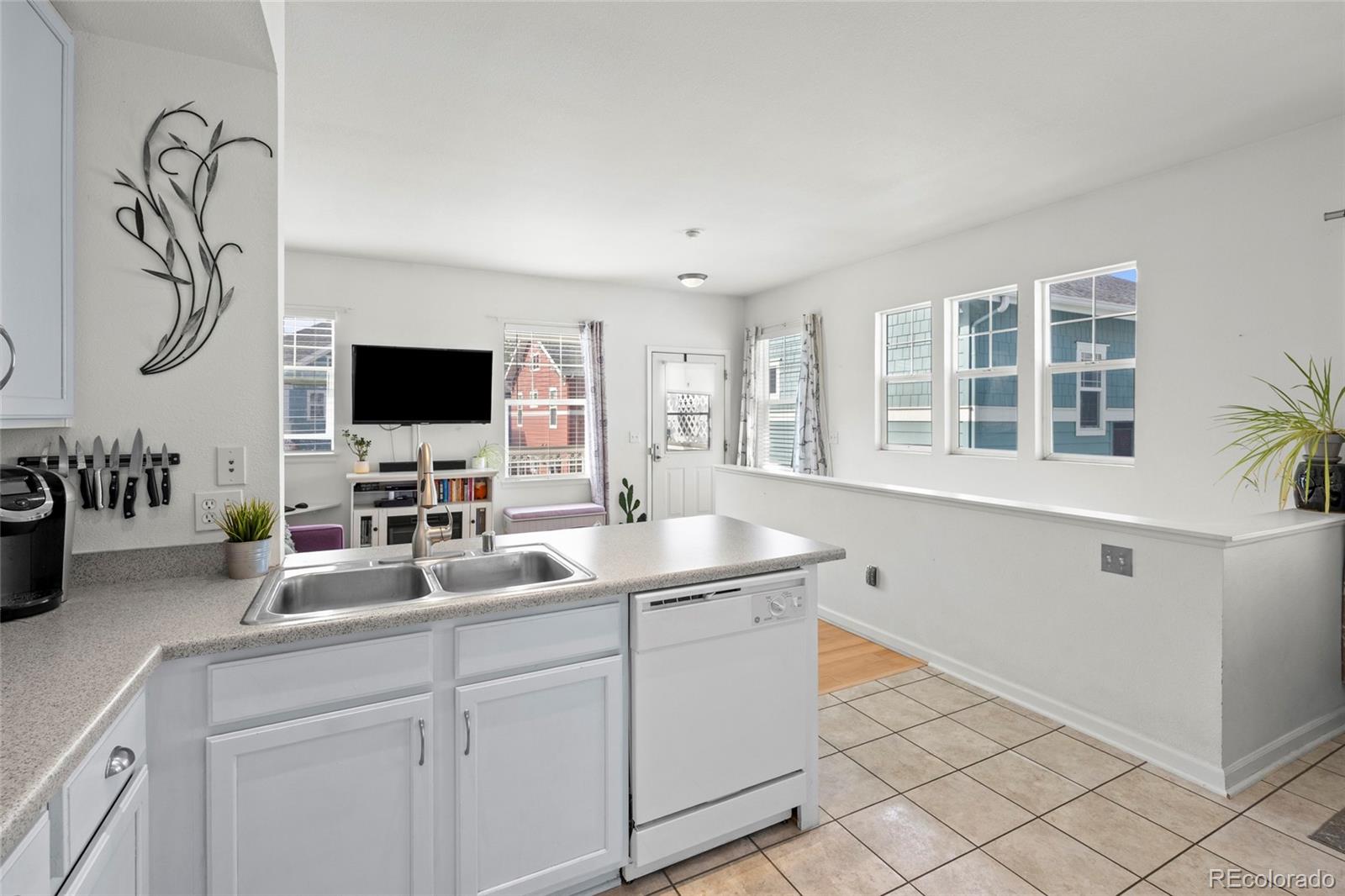 Open-plan kitchen with white cabinets, a double-basin sink, tiled floors, and a view into the living area.
