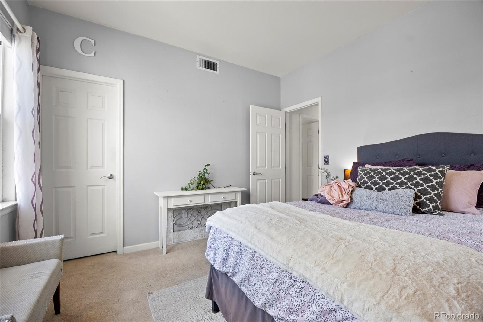 Furnished bedroom with a large lavender-and-white bed, dark upholstered headboard, and window with patterned curtains.