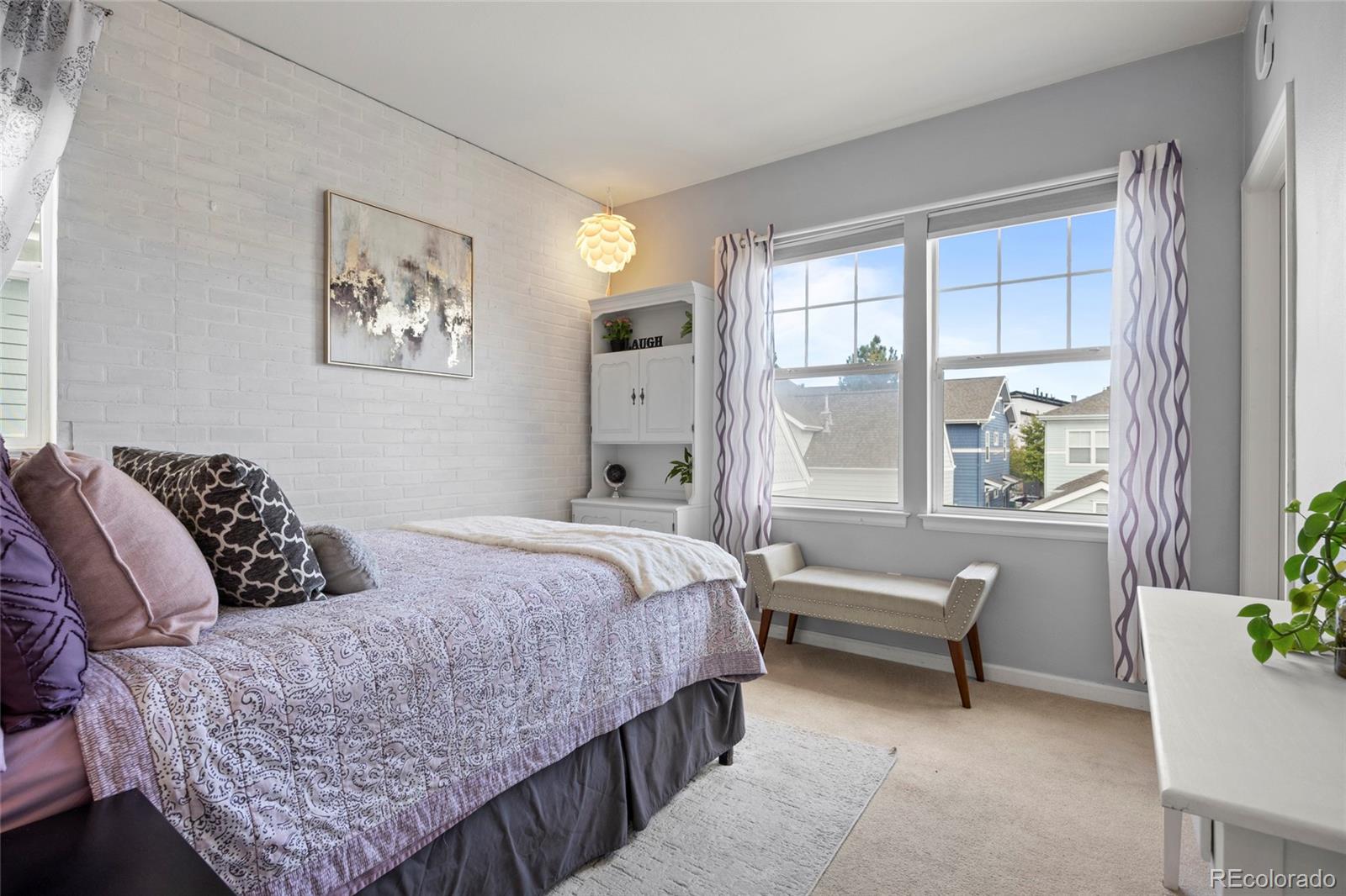 Bedroom with a patterned bed, white brick wall, and large windows overlooking neighboring homes.