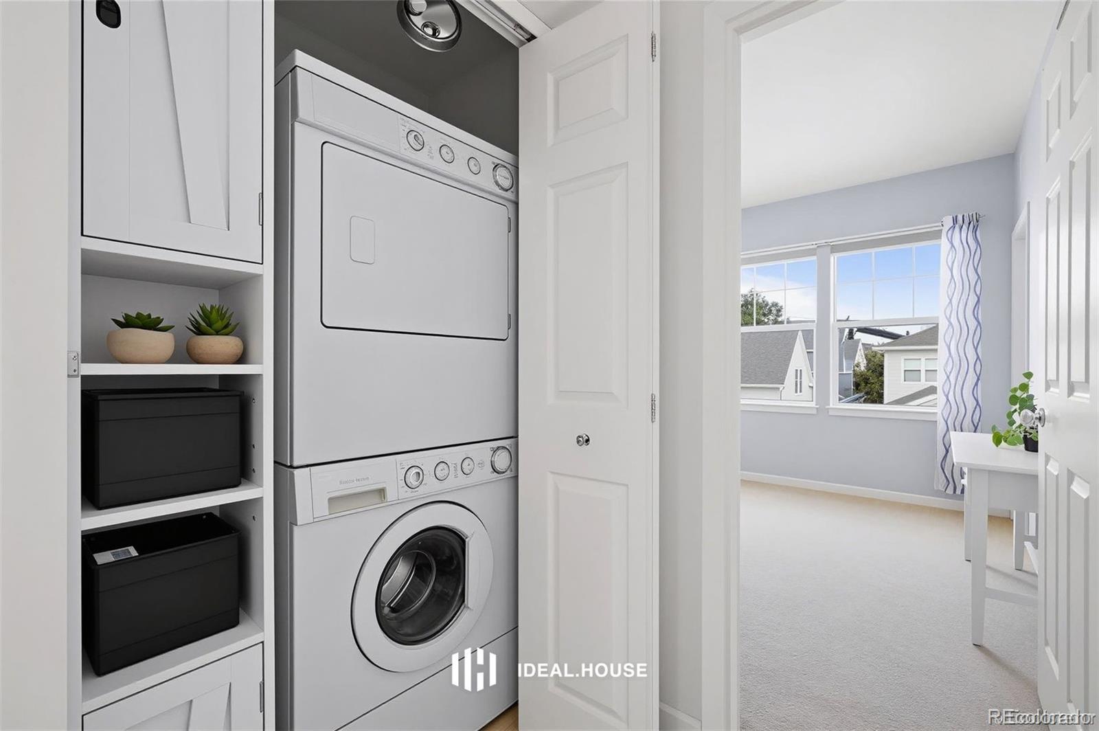 Laundry area with stacked washer and dryer, built-in storage shelves, and an open doorway to a bright room.