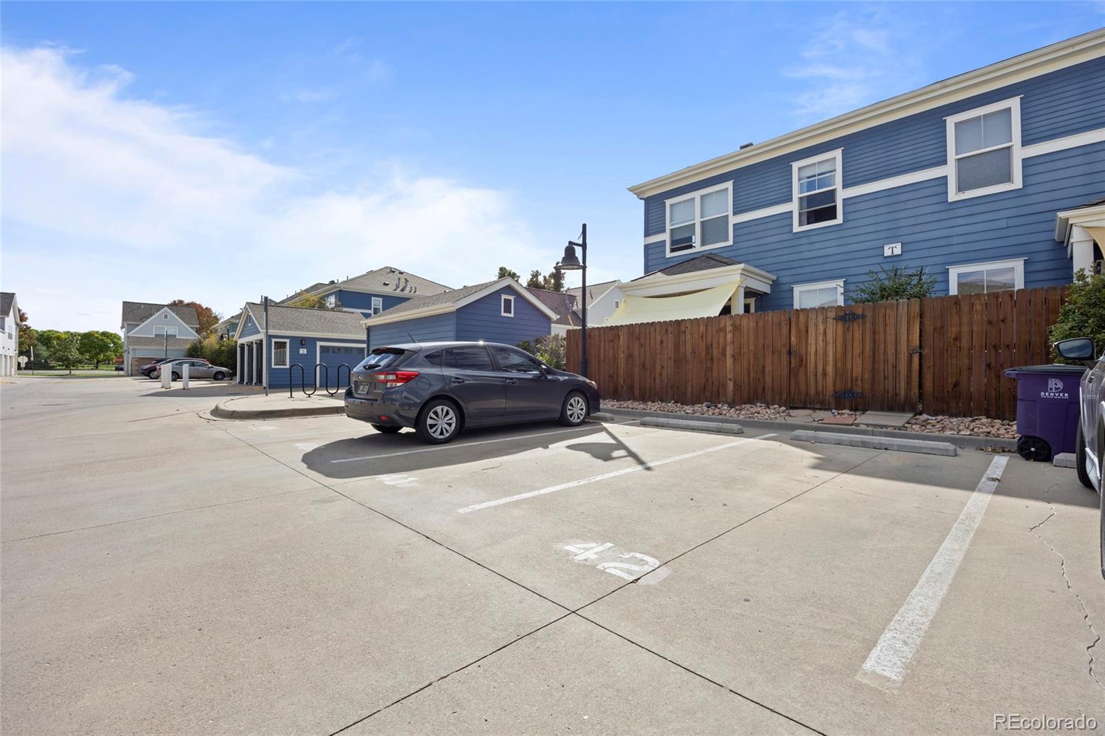 Blue condominium building beside marked parking spaces and a tall wooden fence.