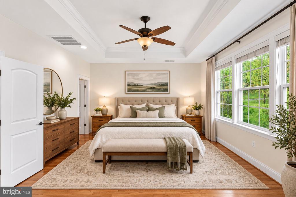 Staged bedroom with a king bed, wood nightstands, and large windows overlooking greenery.