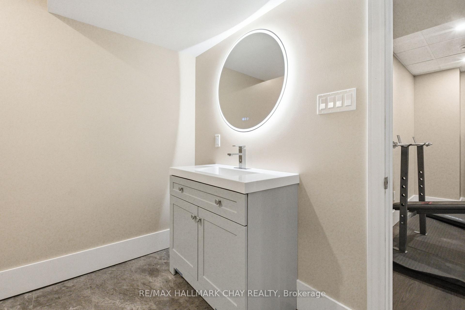 Bathroom vanity with an illuminated round mirror and a view of gym equipment through the doorway.