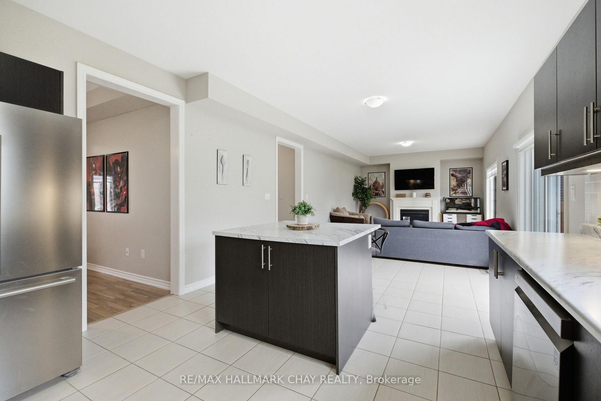 Open kitchen with a dark island and stainless steel refrigerator, overlooking a furnished living room with a fireplace.