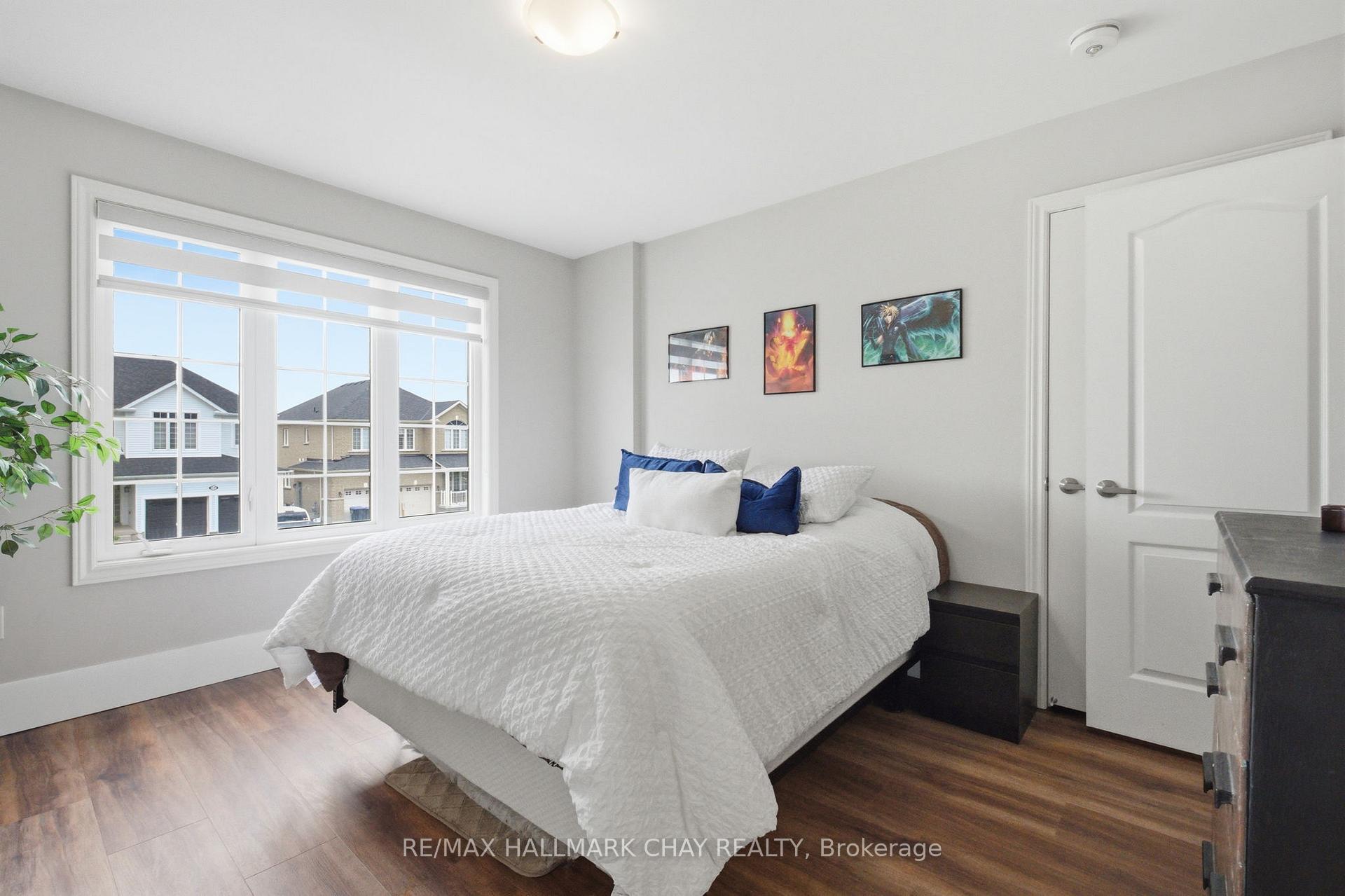 Bright bedroom with a white bed, blue pillows, and a wide window overlooking neighboring homes.