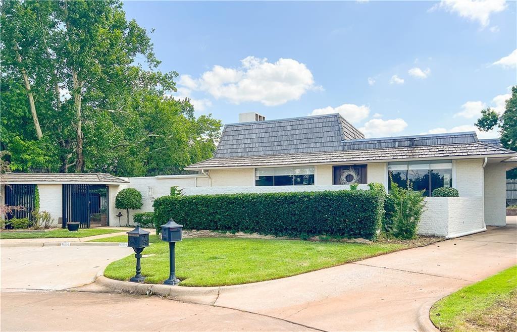 White-brick home with a gray shingle roof, long hedge, curved driveway, and gated entrance.