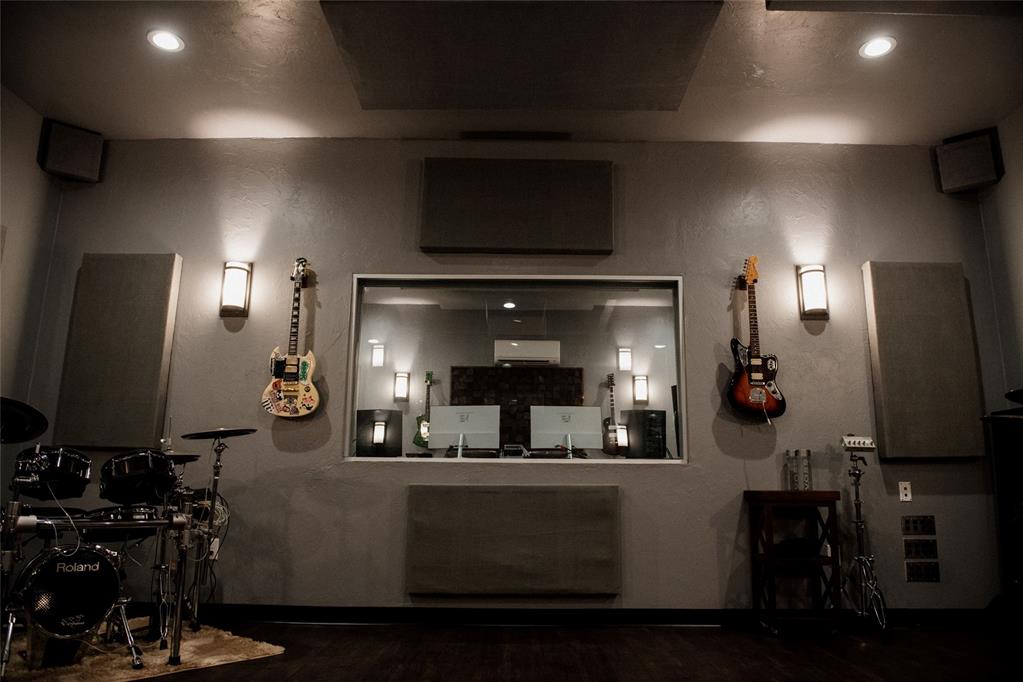 Music studio with acoustic panels, a control-room window, wall-mounted guitars, and an electronic drum kit.