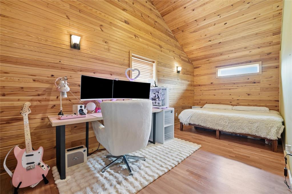Bedroom and workspace with vaulted wood-plank walls, dual-monitor desk, pink guitar, and bed.