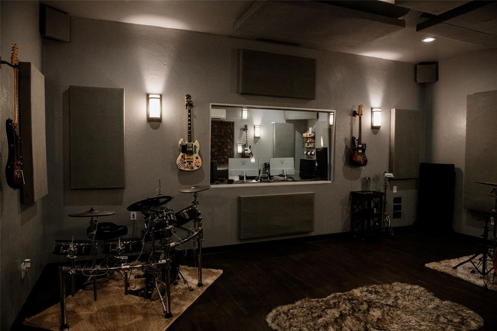 Music room with a drum kit, wall-mounted guitars, acoustic panels, and a recording-room window.