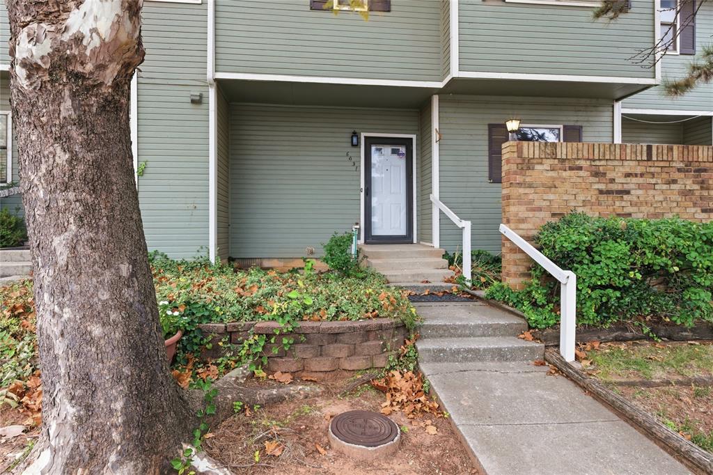 Green-sided home entrance with a white door, concrete walkway, and large tree in the foreground.