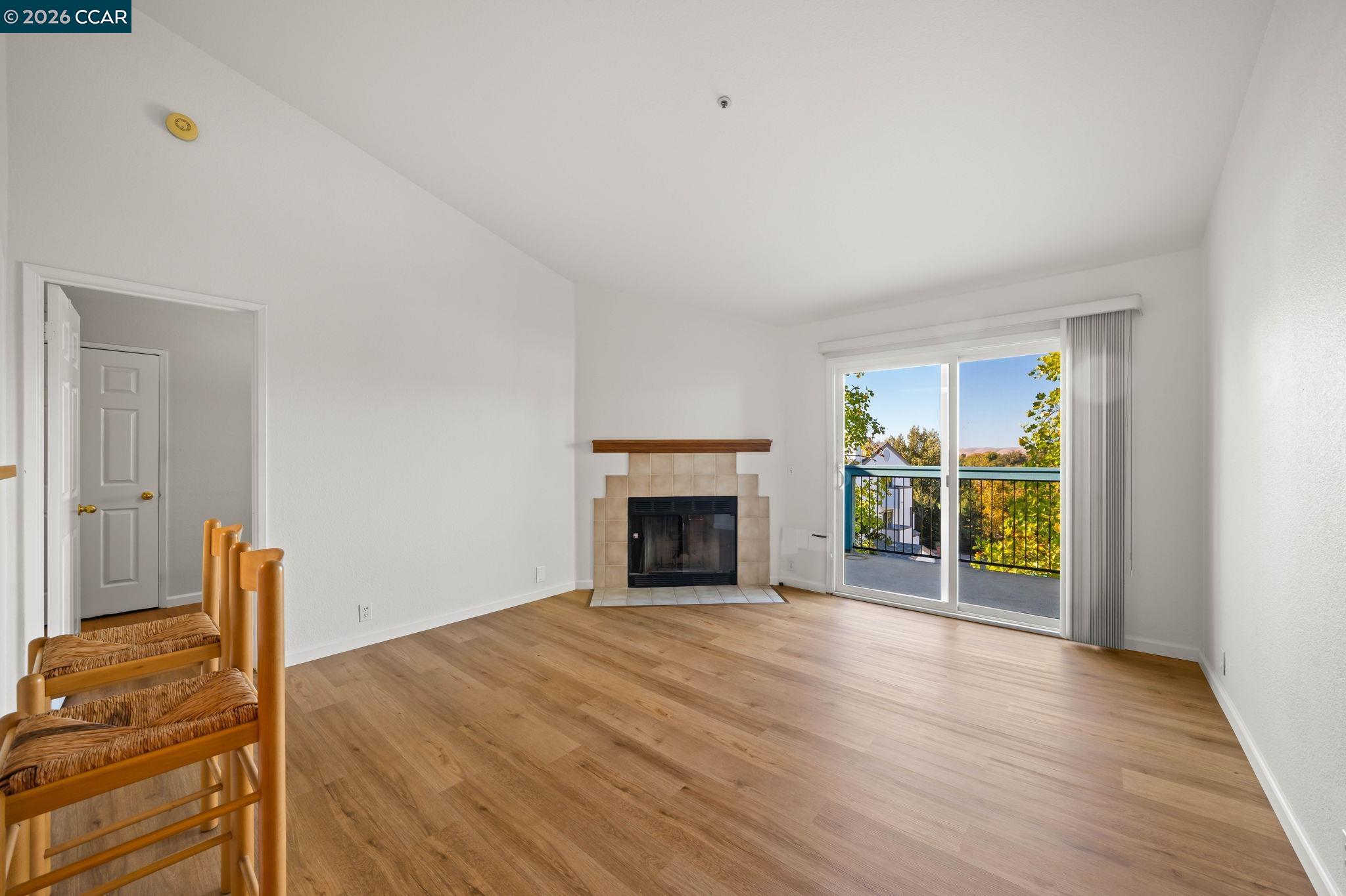Empty living room with a tiled fireplace, wood flooring, and sliding doors to a tree-lined balcony.
