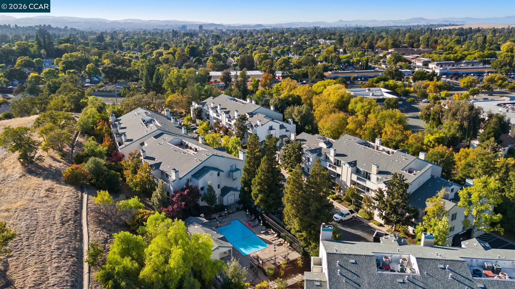 Aerial view of a condominium complex with a swimming pool, leafy trees, and distant hills.