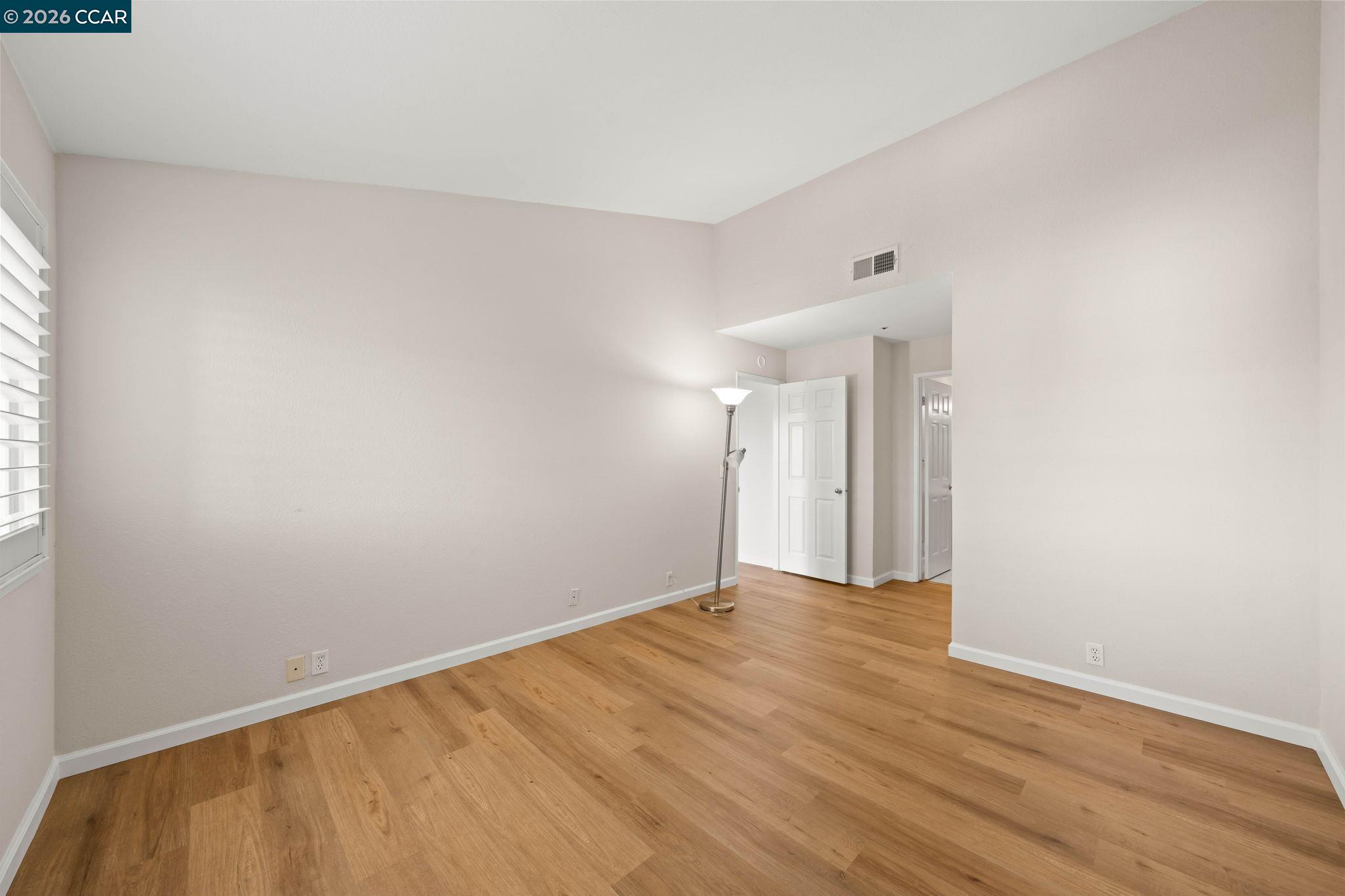 Empty living room with light wood floors, pale walls, and a floor lamp beside the open doorway.